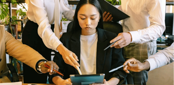 Une jeune femme en blazer noir qui semble dépassée et confuse tout en tenant une tablette, alors que les mains de ses collègues pointent des stylos dans des directions contradictoires vers elle et l'écran.